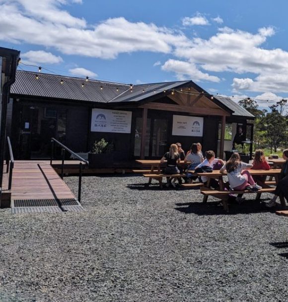 People sit at wooden picnic tables outside a modern, dark-colored building with large windows under a bright blue sky with scattered clouds. String lights hang above the entrance. The area is gravel-covered.