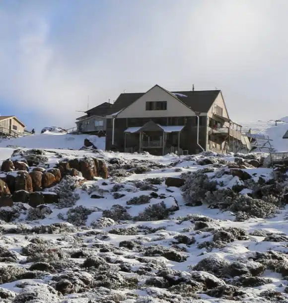 A large house with a sloped roof sits on a rocky, snow-dusted hillside under a partly cloudy sky. Sparse vegetation and patches of snow cover the ground in front of the building.
