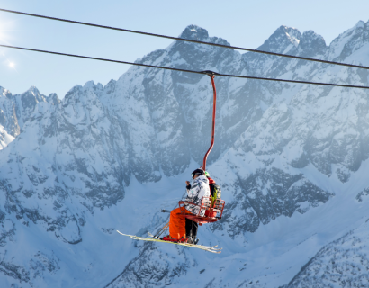 Two skiers wearing winter gear sit on a red ski lift ascending with snow-covered mountains and a clear sky in the background.