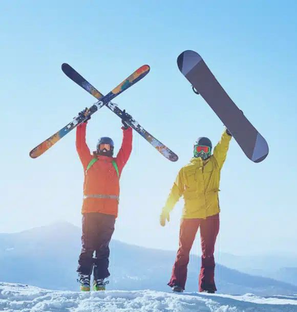 Two people in winter gear stand on snowy ground; one holds skis crossed above their head, the other lifts a snowboard. Mountains and a clear blue sky are in the background.