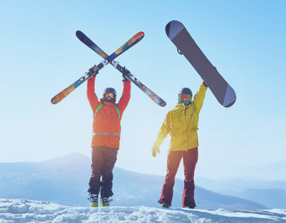 Two people in winter gear stand on a snowy slope; one holds skis crossed overhead, the other raises a snowboard. Mountains and blue sky are in the background.