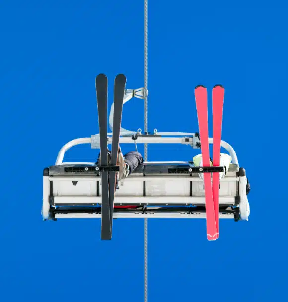 A ski lift chair viewed from below against a clear blue sky, carrying two people with one pair of black skis and one pair of red skis attached to the back.