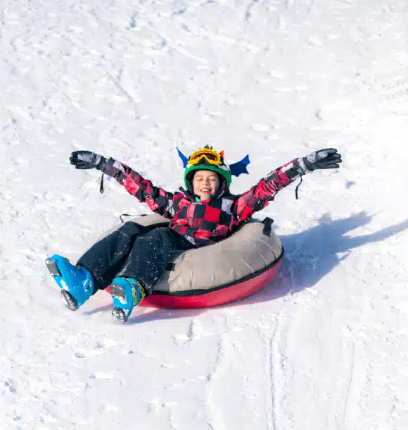 A child wearing a helmet and gloves smiles with arms outstretched while sliding down a snowy hill on an inflatable snow tube.