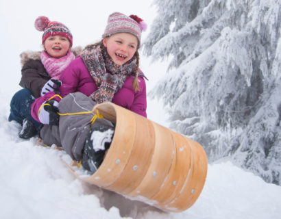 Two smiling children in winter clothes ride a wooden sled down a snowy hill, surrounded by snow-covered trees and laughing joyfully.