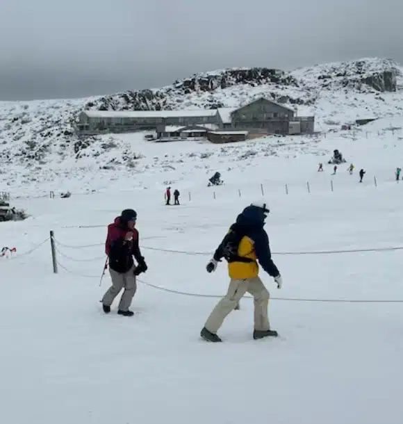 Two people wearing winter clothing walk on a snowy landscape with a few others in the background. A building and rocky hillside covered in snow are visible under an overcast sky.